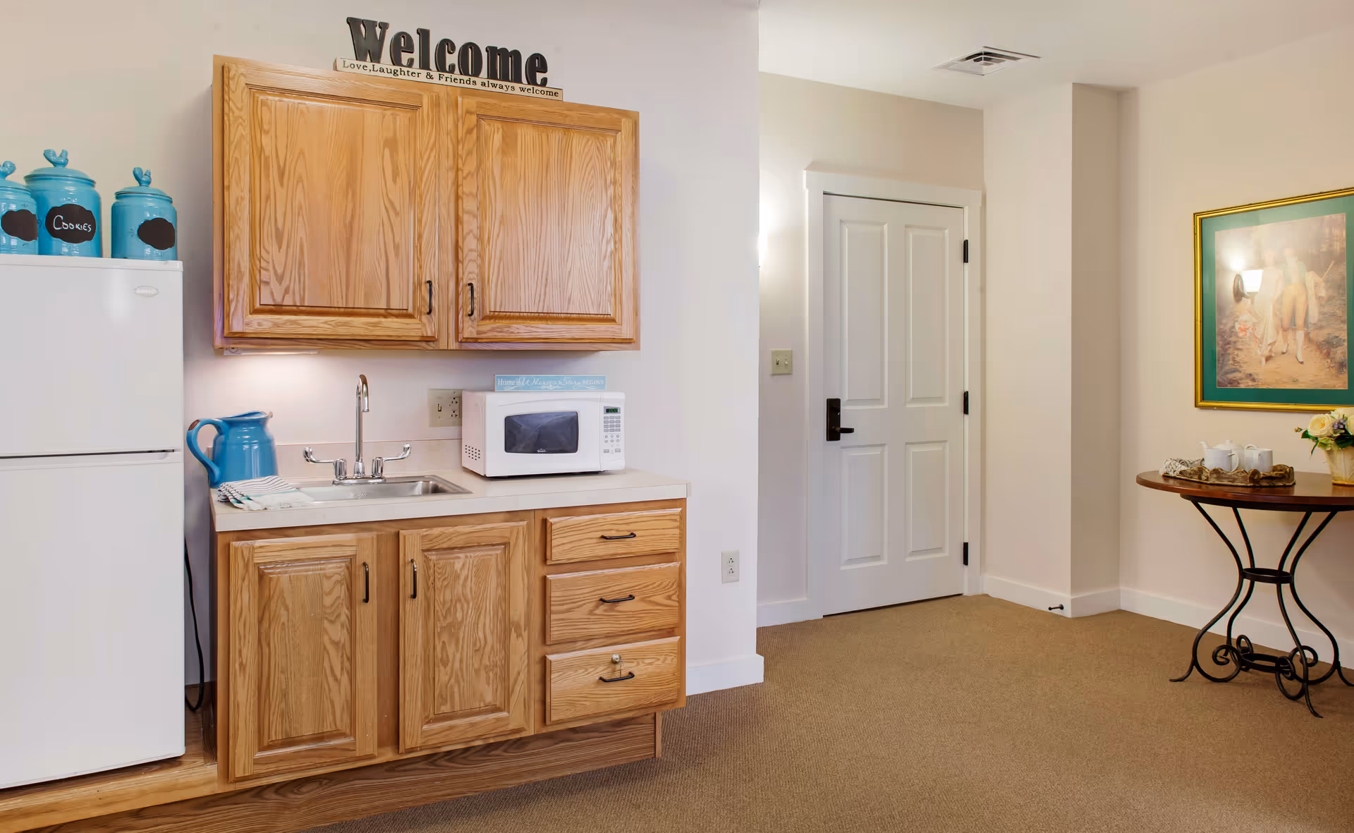 A small kitchenette area with wooden cabinets, a white microwave on the countertop, a stainless steel sink, and a white refrigerator with blue canisters labeled 'Cookies' on top. To the right, there is a white door and a round wooden table with a tea set and a vase of flowers, along with a framed painting on the wall.