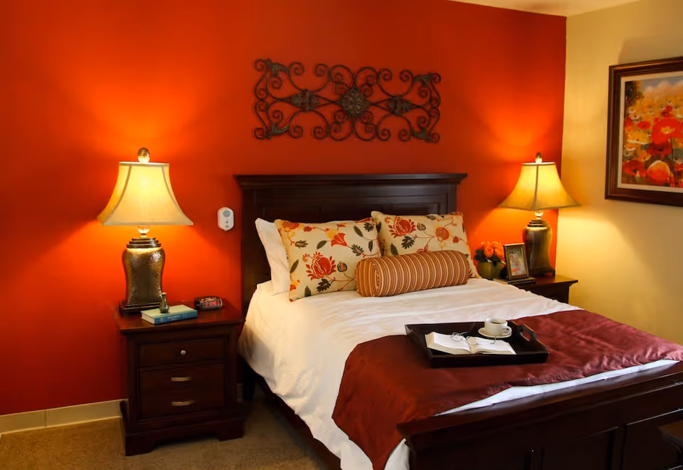 A cozy bedroom with a dark wooden bed frame and white bedding accented by floral and striped pillows. There are two bedside tables, each with a lamp, and a decorative metal wall art piece above the bed. A tray with a cup, glasses, and a book is placed on the bed. The walls are painted red and beige, and a colorful floral painting hangs on the beige wall.