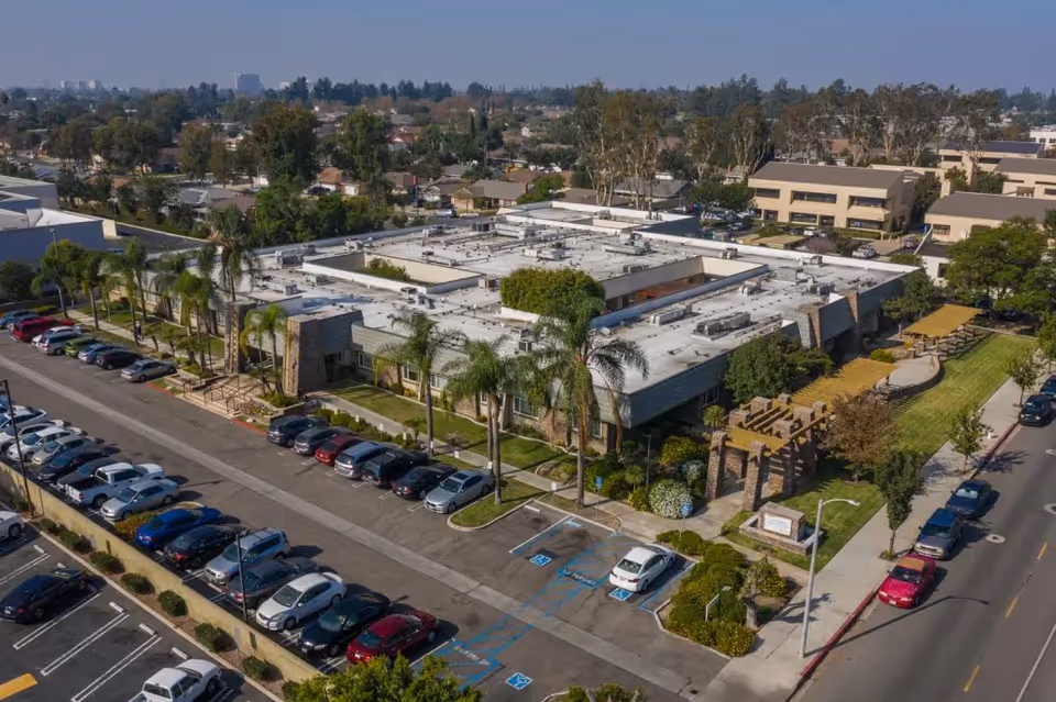 Aerial view of a large single-story building with a flat roof surrounded by palm trees and greenery. The building is situated in a suburban area with parked cars along the street and parking lot adjacent to it. There is a pergola structure and a small grassy area next to the building. Residential houses and other buildings are visible in the background under a clear sky.