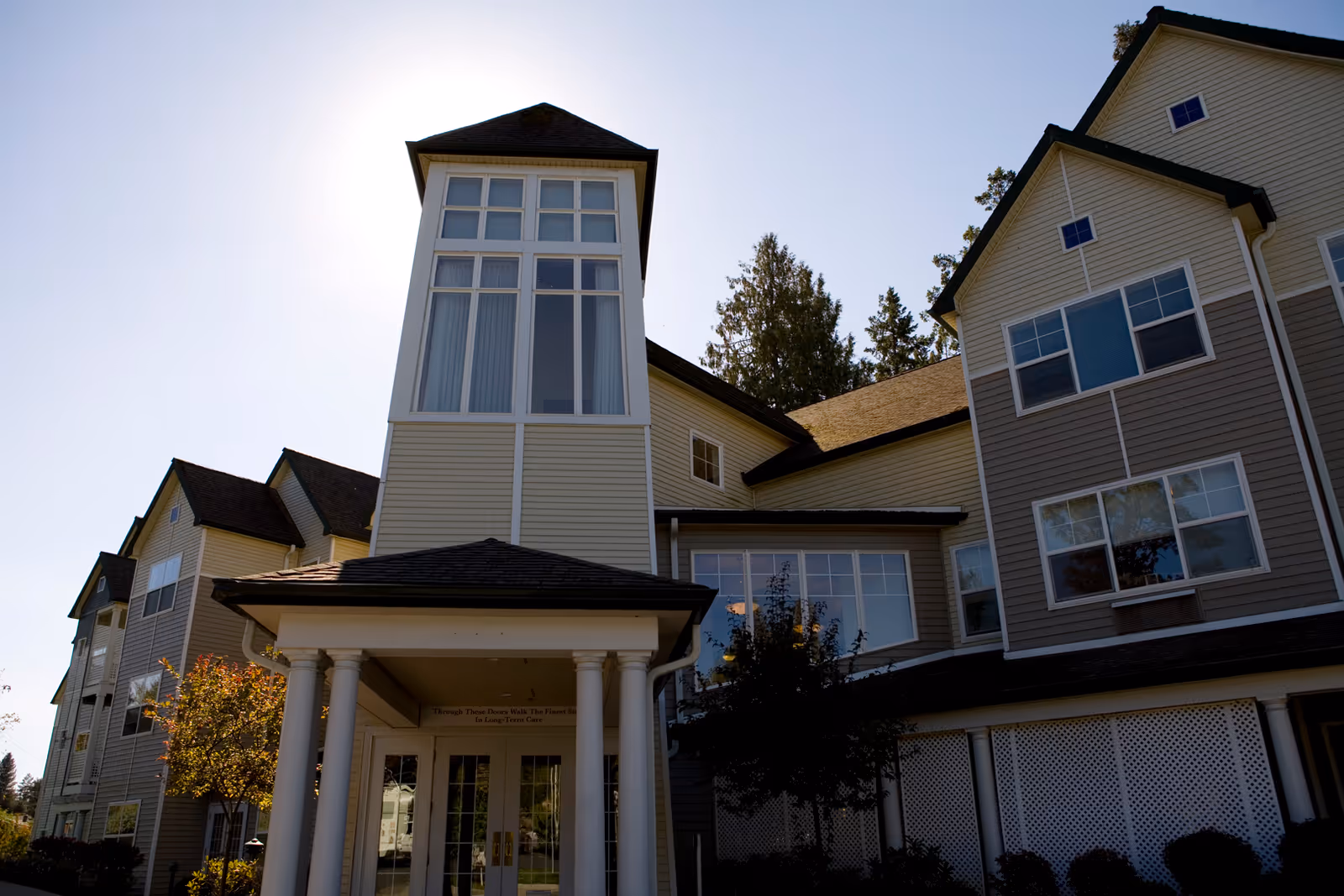 Front exterior of a multi-story beige senior living building with a tall central tower and a columned entrance portico.