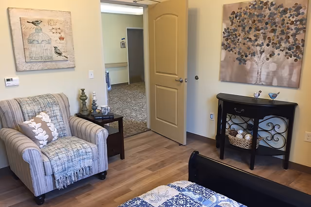 A cozy bedroom corner in a senior living facility with a striped armchair draped with a blue throw and a decorative pillow. Next to the chair is a small wooden side table with candles and a cup. The room has light wood flooring and beige walls adorned with two paintings: one of birds and a birdcage, and another of a stylized tree. A black console table with decorative items and a basket is against the wall. The door is open, showing a carpeted hallway outside.