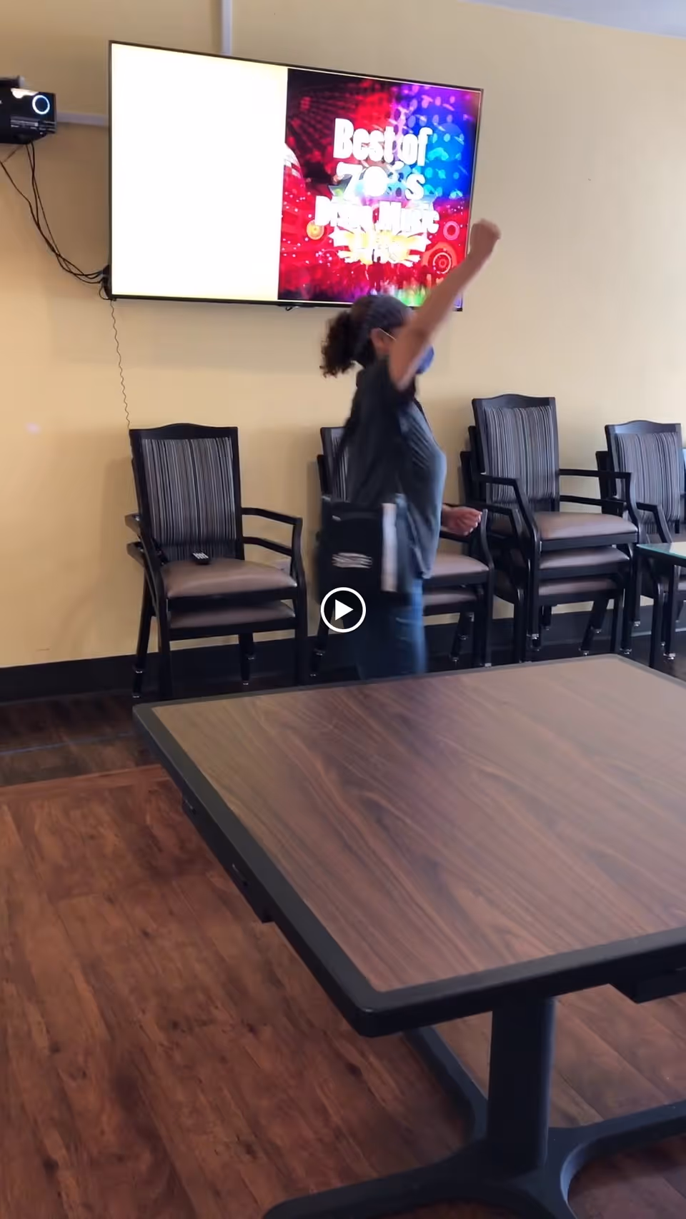 A person wearing a mask and dark clothing stands in a room with wooden flooring and several stacked chairs with striped cushions against a beige wall. A large flat-screen TV mounted on the wall displays a colorful screen with the text 'Best of 70's Karaoke'. A wooden table is in the foreground.