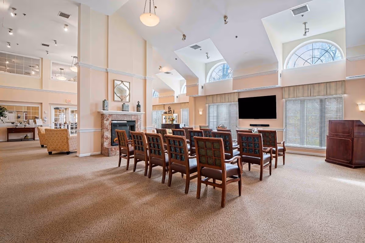 A spacious, well-lit common area with rows of wooden chairs facing a large flat-screen TV mounted on the wall. The room features high ceilings with arched windows letting in natural light, a fireplace with decorative items on the mantel, and a podium to the right. The carpeted floor and soft beige walls create a warm and inviting atmosphere.