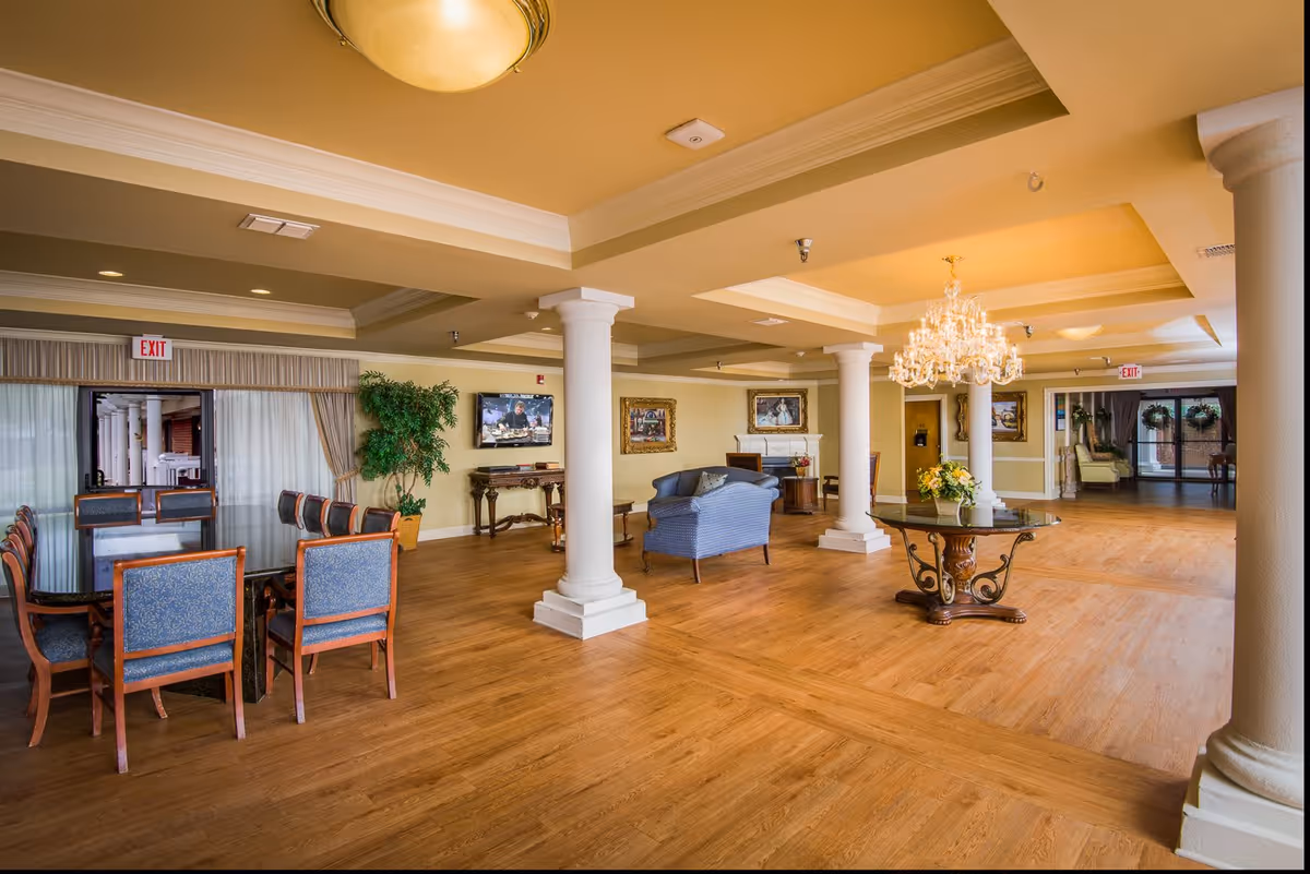 Spacious and well-lit interior common area of The Gables At Spring Lake Assisted Living featuring wooden flooring, white columns, a chandelier above a round glass table with a floral centerpiece, a seating area with blue upholstered chairs and a sofa, framed paintings on the walls, a flat-screen TV, and a dining table with multiple chairs near large windows with curtains.
