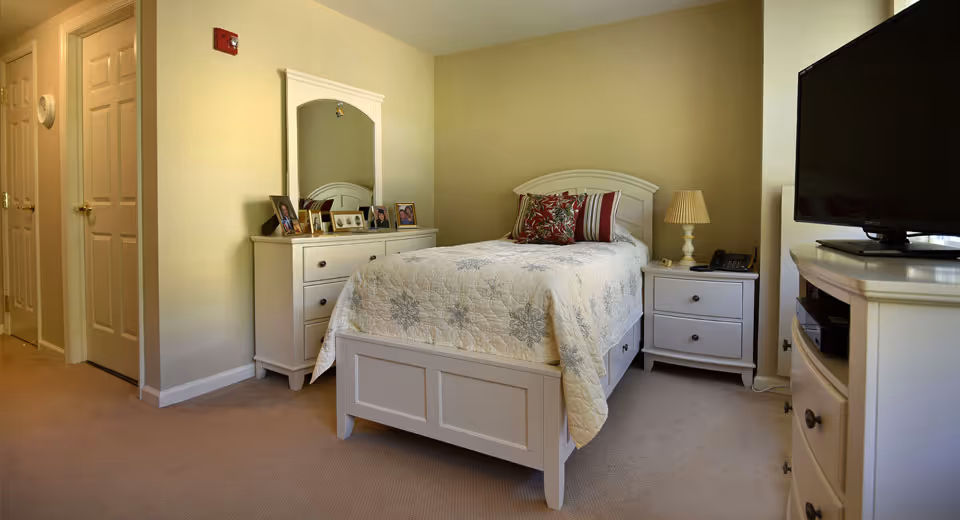 A cozy bedroom with a single bed covered in a white quilt with floral patterns. The bed has decorative pillows and is flanked by a white nightstand with a lamp and a telephone. Across from the bed is a white dresser with a flat-screen TV on top. Another white dresser with a mirror and framed photos is positioned against the wall. The room has beige walls and carpeted flooring.