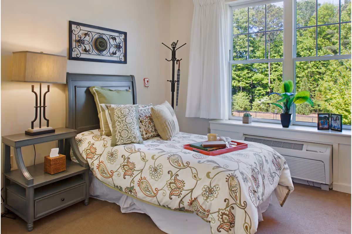 A bright, neatly made bedroom with a patterned single bed, bedside table and lamp, coat rack, and a large window overlooking trees.