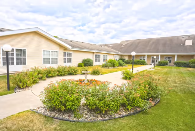 Courtyard with a circular flower bed, paved walkways, lamp posts, and a single-story beige building surrounding a grassy lawn.