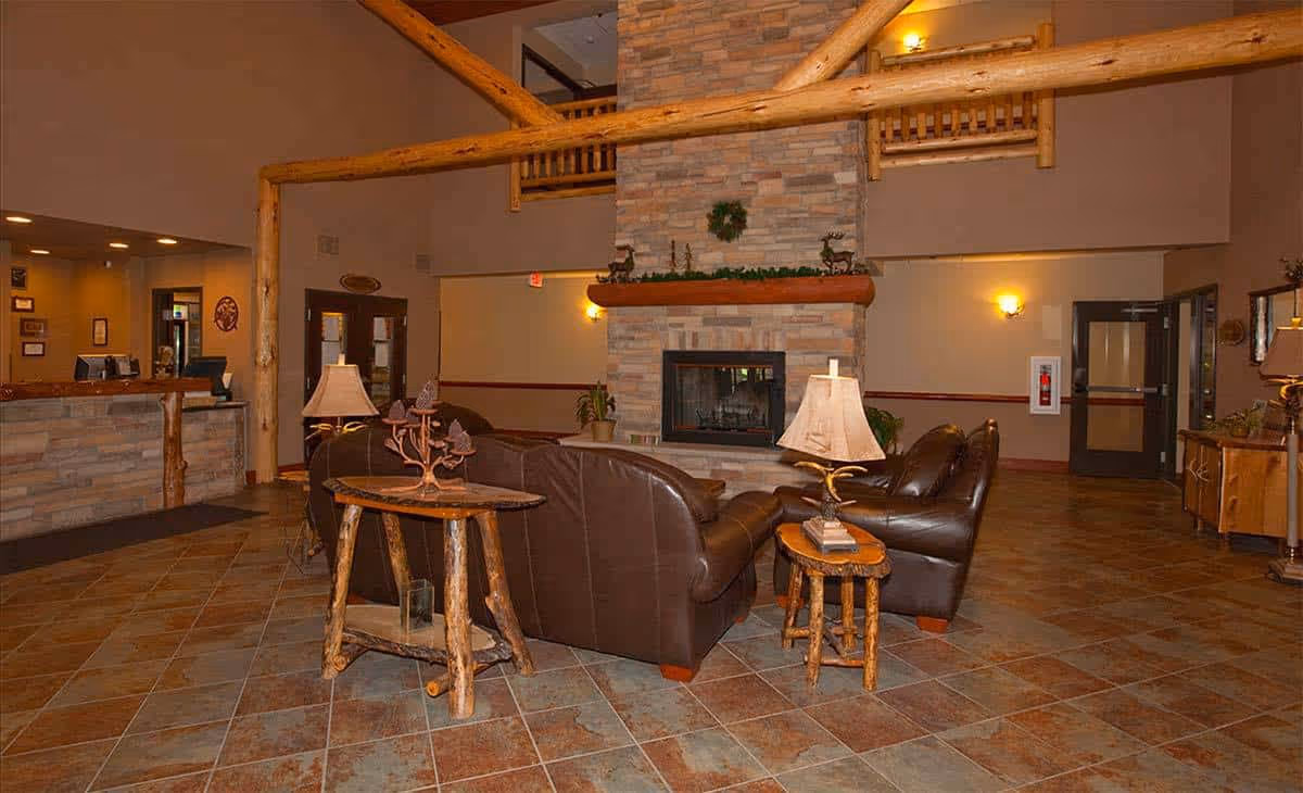 A cozy senior living facility common area with brown leather sofas arranged around a stone fireplace. The room features rustic wooden furniture including side tables with lamps, a tiled floor, and warm lighting. The reception desk is visible to the left, and there are doors and wall sconces along the walls.