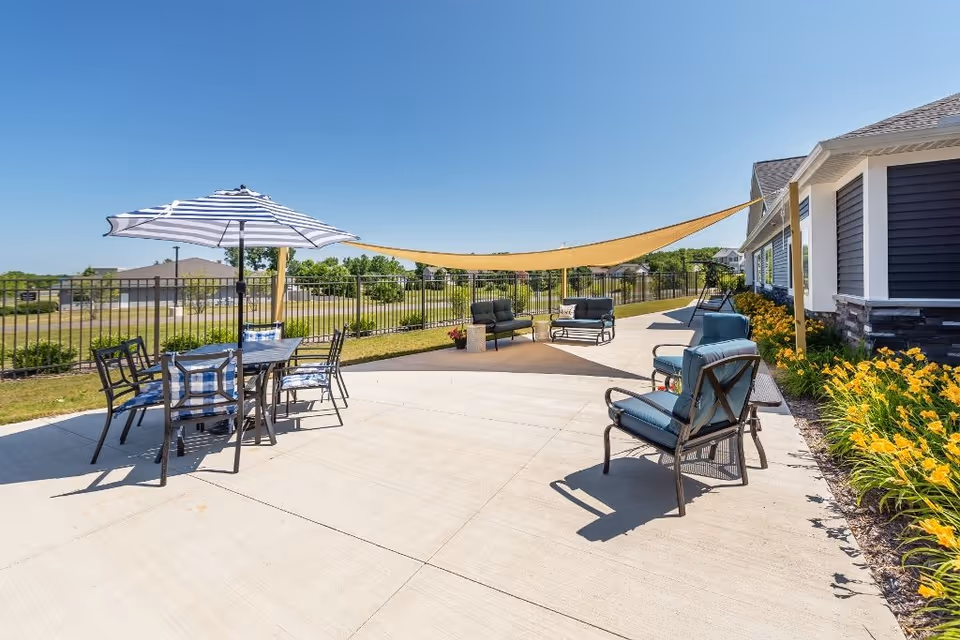 Outdoor patio area with several seating arrangements including a table with chairs under a striped umbrella, cushioned chairs, and a shaded area with a large beige sunshade. The patio is adjacent to a building with dark siding and stone accents, surrounded by a black metal fence and landscaped with yellow flowers. The sky is clear and blue.