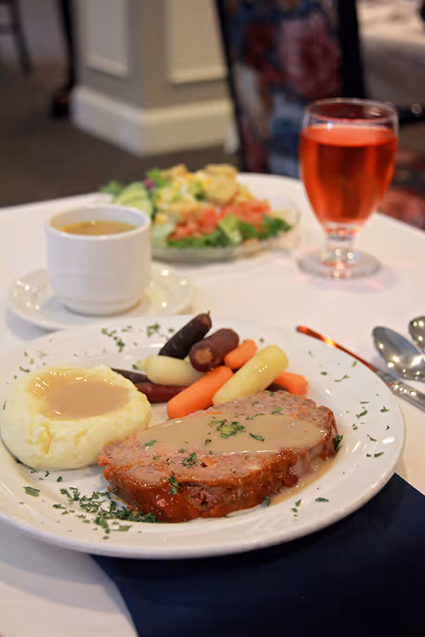 A plated meal consisting of meatloaf with gravy, mashed potatoes with gravy, and a mix of baby carrots and other vegetables on a white plate garnished with herbs. In the background, there is a cup of soup, a glass of red beverage, and a salad plate on a white tablecloth in a dining setting.