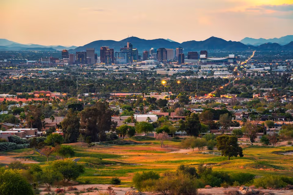 View of a city skyline at sunset with mountains in the background and a green park area with trees and houses in the foreground.