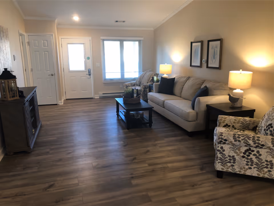 A well-lit living room with wooden flooring, a beige sofa with dark cushions, two patterned armchairs, two side tables with lamps, a coffee table with decorative items, a window with blinds, and a white front door with a small window. The walls are light-colored and there are two framed pictures hanging above the sofa.