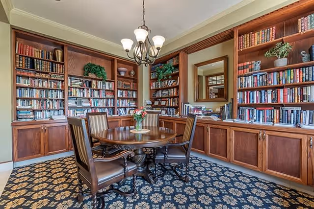 A cozy library room with wooden bookshelves filled with books lining the walls. In the center, there is a round wooden table with four upholstered chairs around it. A chandelier with multiple lights hangs above the table. The room has a patterned carpet and a large mirror on one wall reflecting part of the room.