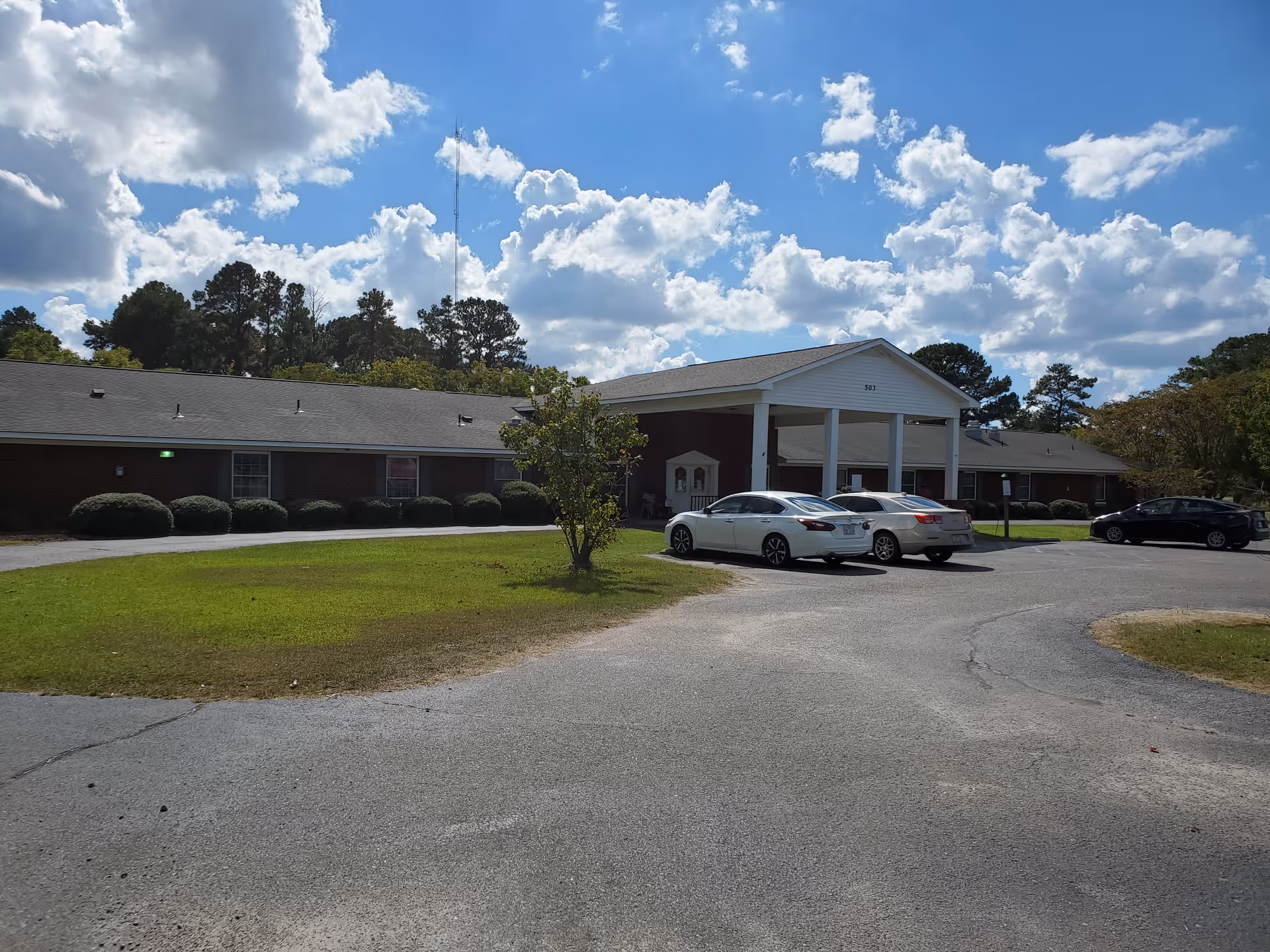 Exterior view of a single-story brick building with a covered entrance supported by white columns. There are three cars parked in front of the building on a paved driveway, with a grassy area and trees surrounding the property under a partly cloudy blue sky.