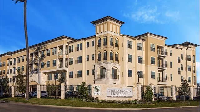 Exterior view of a large, multi-story senior living facility building with beige and light brown walls, multiple balconies, and a sign in front that reads 'The Solana Preserve Vintage Park'. The sky is clear and blue.
