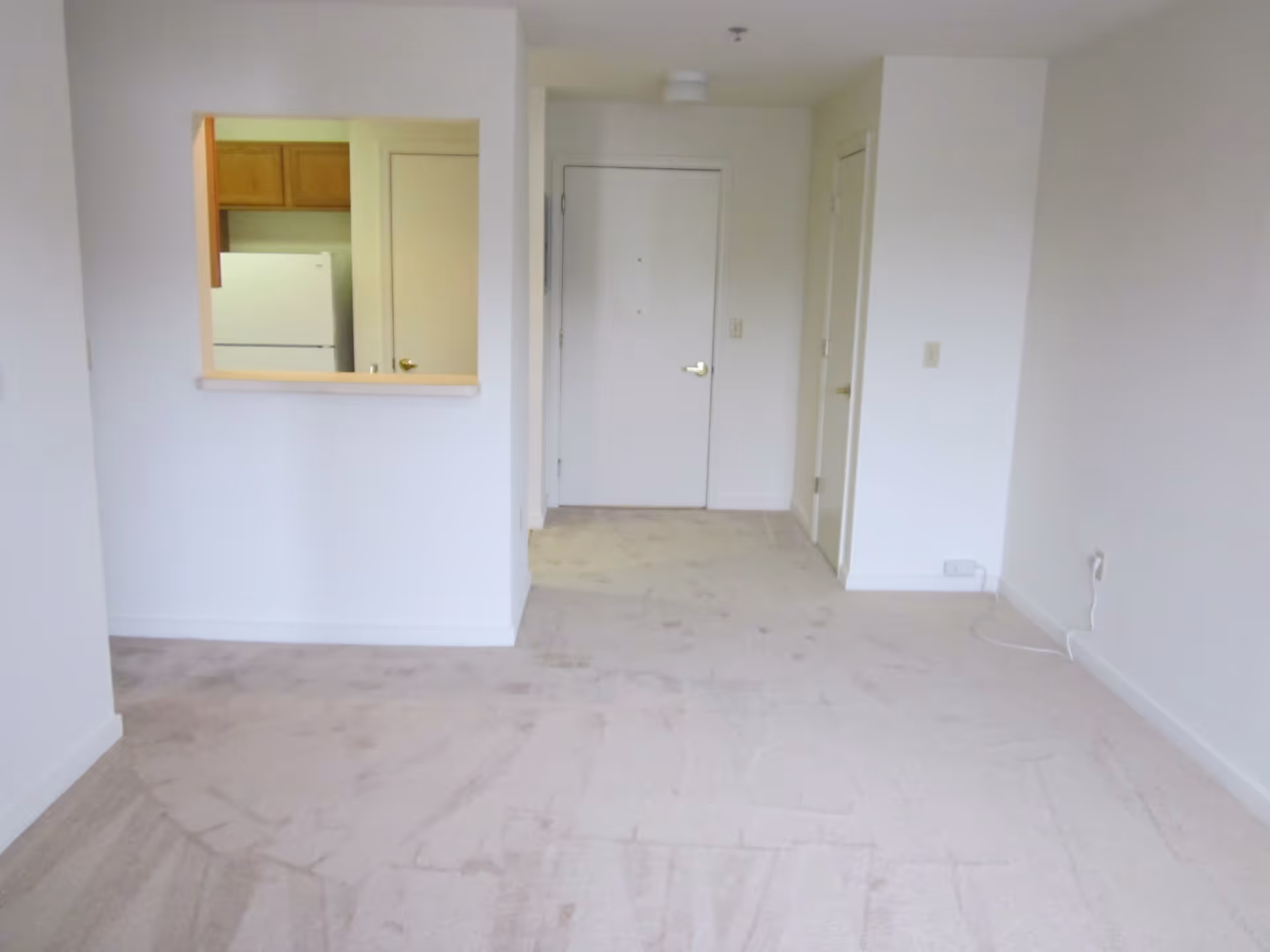 Empty room with beige carpet flooring and white walls. There is a white door at the far end, a small kitchen window opening on the left showing a refrigerator and wooden cabinets, and a closed door to the right of the main door. An electrical outlet with a plugged-in white cable is visible on the right wall.