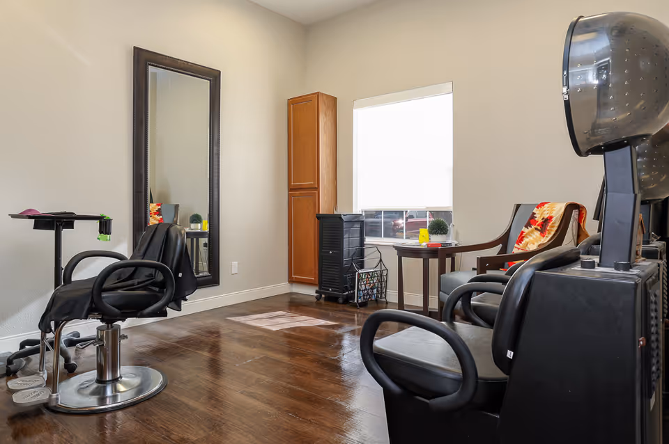 Interior of a salon room with a black salon chair in front of a large wall mirror, a wooden cabinet, a window with natural light, and two black hair dryer chairs with a small round table between them holding a small plant and a book.