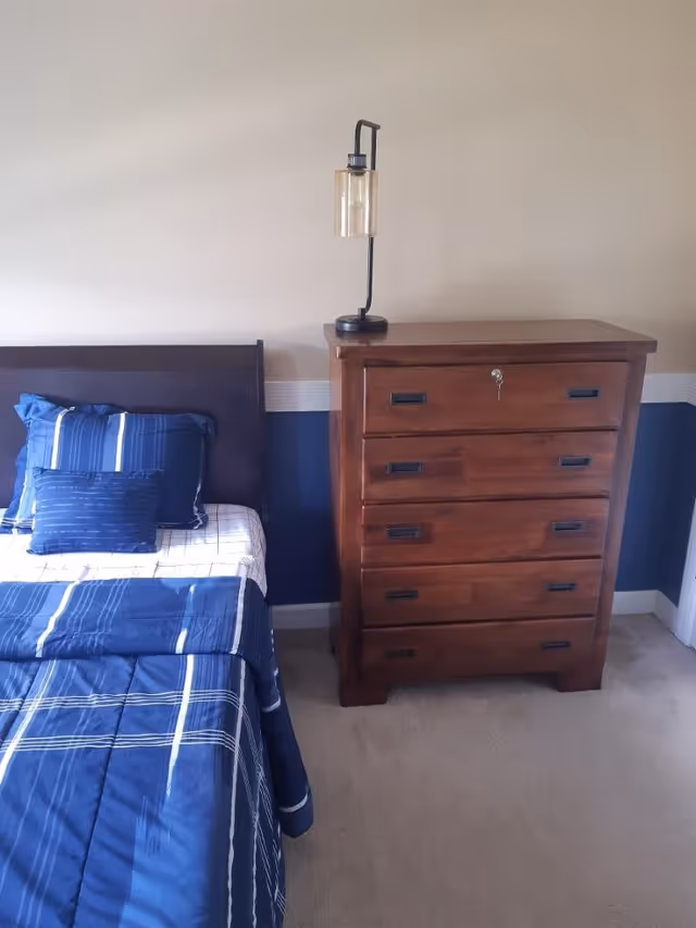 A bedroom corner featuring a bed with blue and white striped bedding and pillows, next to a wooden chest of drawers with a modern lamp on top. The walls are painted beige with a blue lower section and white trim.