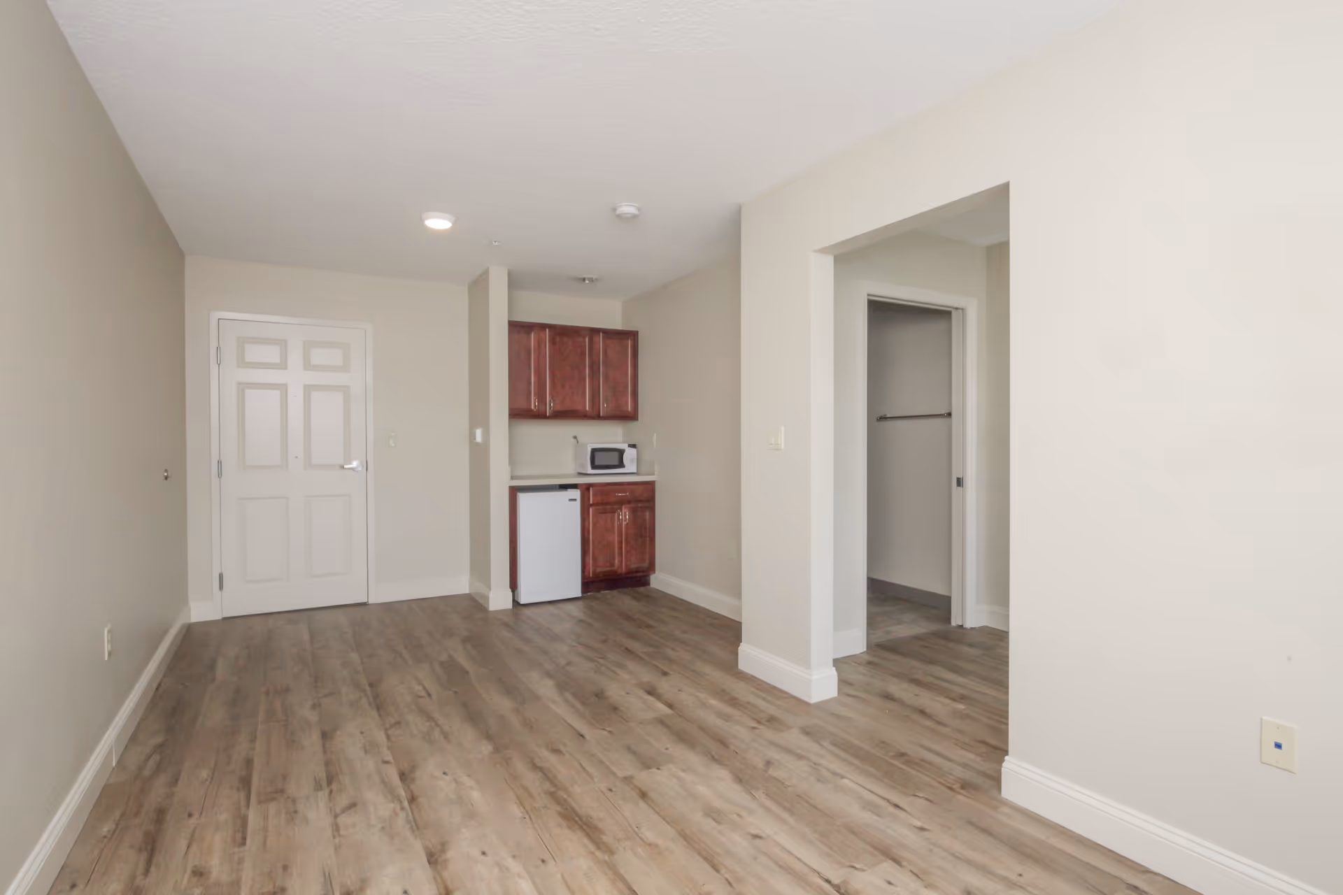 Empty room with light beige walls and wood-look flooring. There is a white door on the left side, a small kitchenette area with dark wood cabinets, a microwave, and a mini fridge in the center background. To the right, there is an open doorway leading to another room with a towel rack visible.