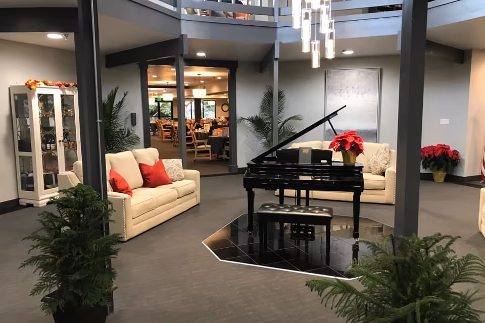 A spacious senior living facility common area with a black grand piano on a tiled platform in the center, surrounded by two beige sofas with red and beige cushions. The room features modern lighting fixtures hanging from the ceiling, potted plants, and a view into a dining area with tables and chairs in the background.