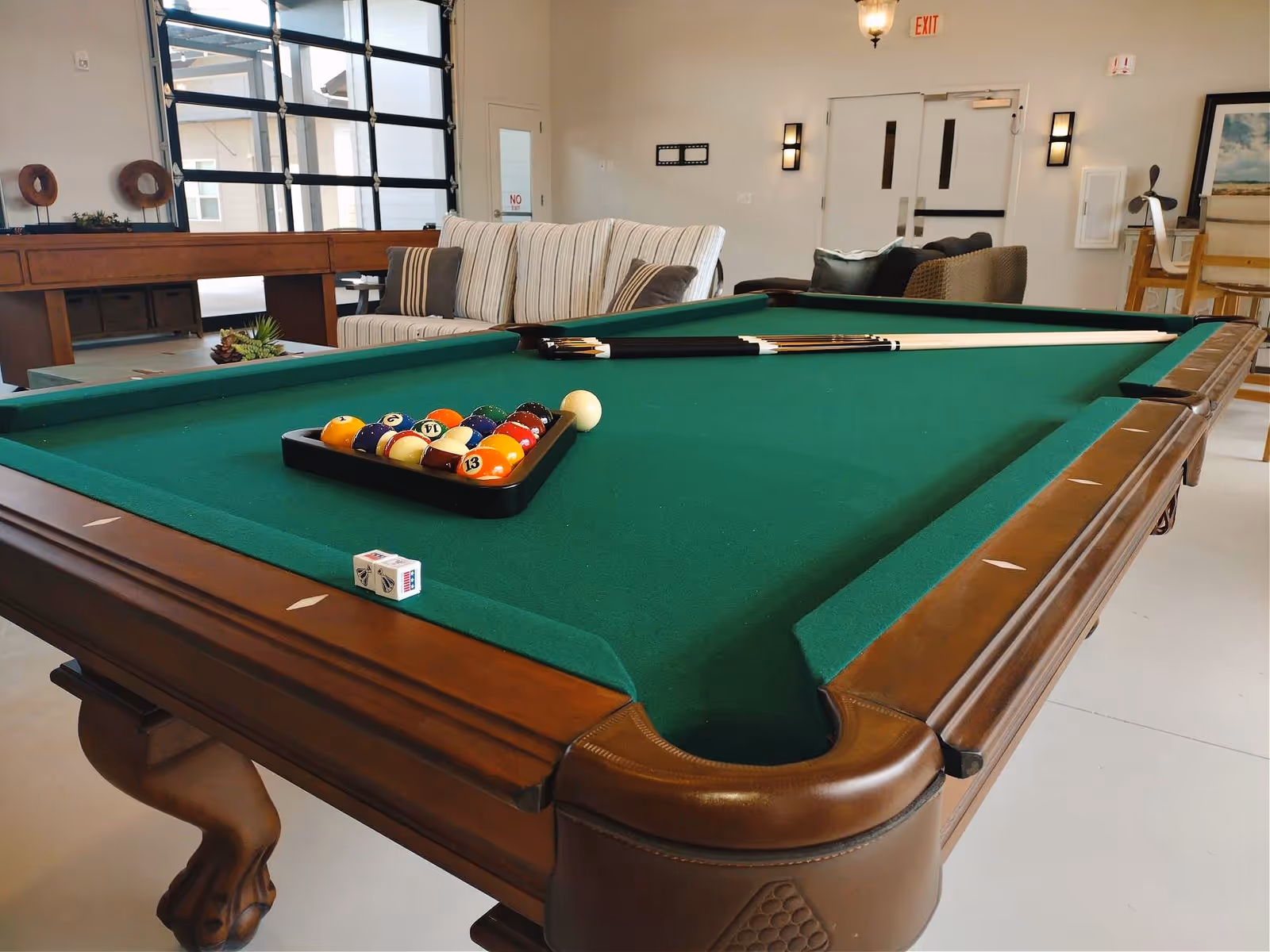 A pool table with a green felt surface set up for a game with billiard balls racked in a triangle and two pool cues resting on the table. In the background, there is a striped sofa, a shuffleboard table, and other seating furniture in a well-lit recreational room with large windows and a door marked with an exit sign.