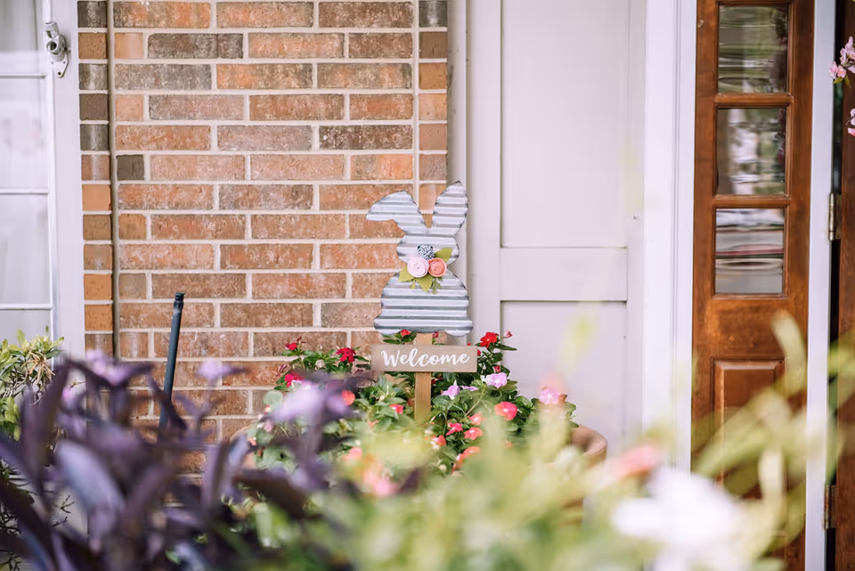 Front porch with potted flowers and a decorative bunny "Welcome" sign against a brick wall beside a wooden door.