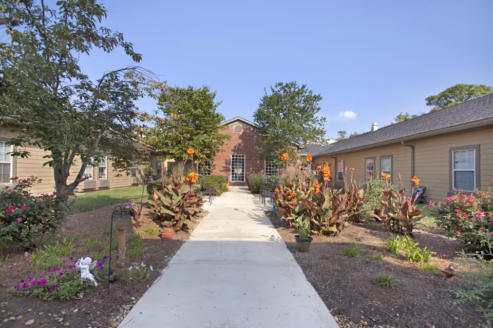 A paved walkway leading through a garden area with various plants and flowers, flanked by two single-story buildings with beige siding and multiple windows, under a clear blue sky.