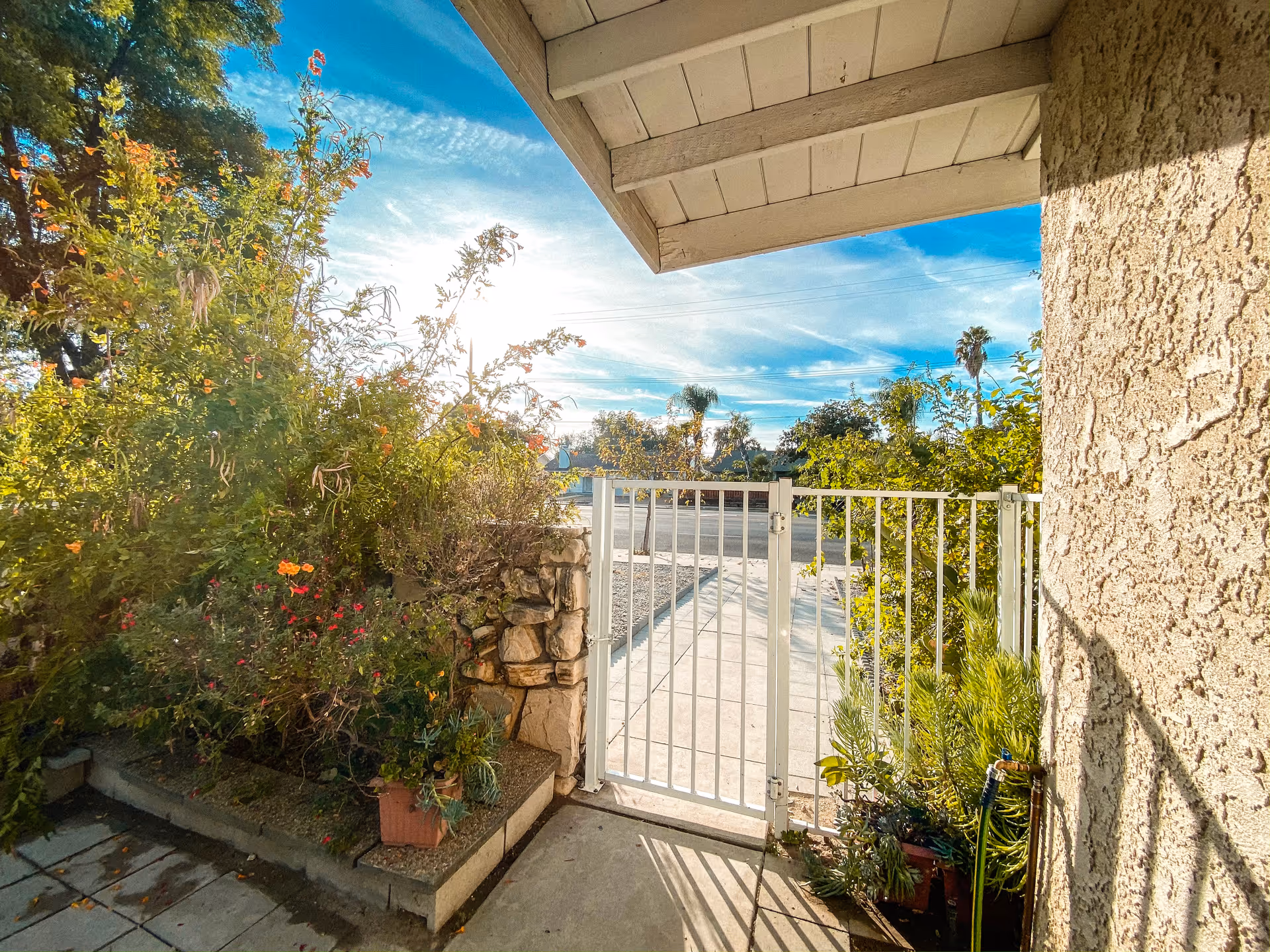 View of a gated entrance from a covered porch area with plants and greenery on the left side, a stone wall, and a clear blue sky with some clouds in the background.