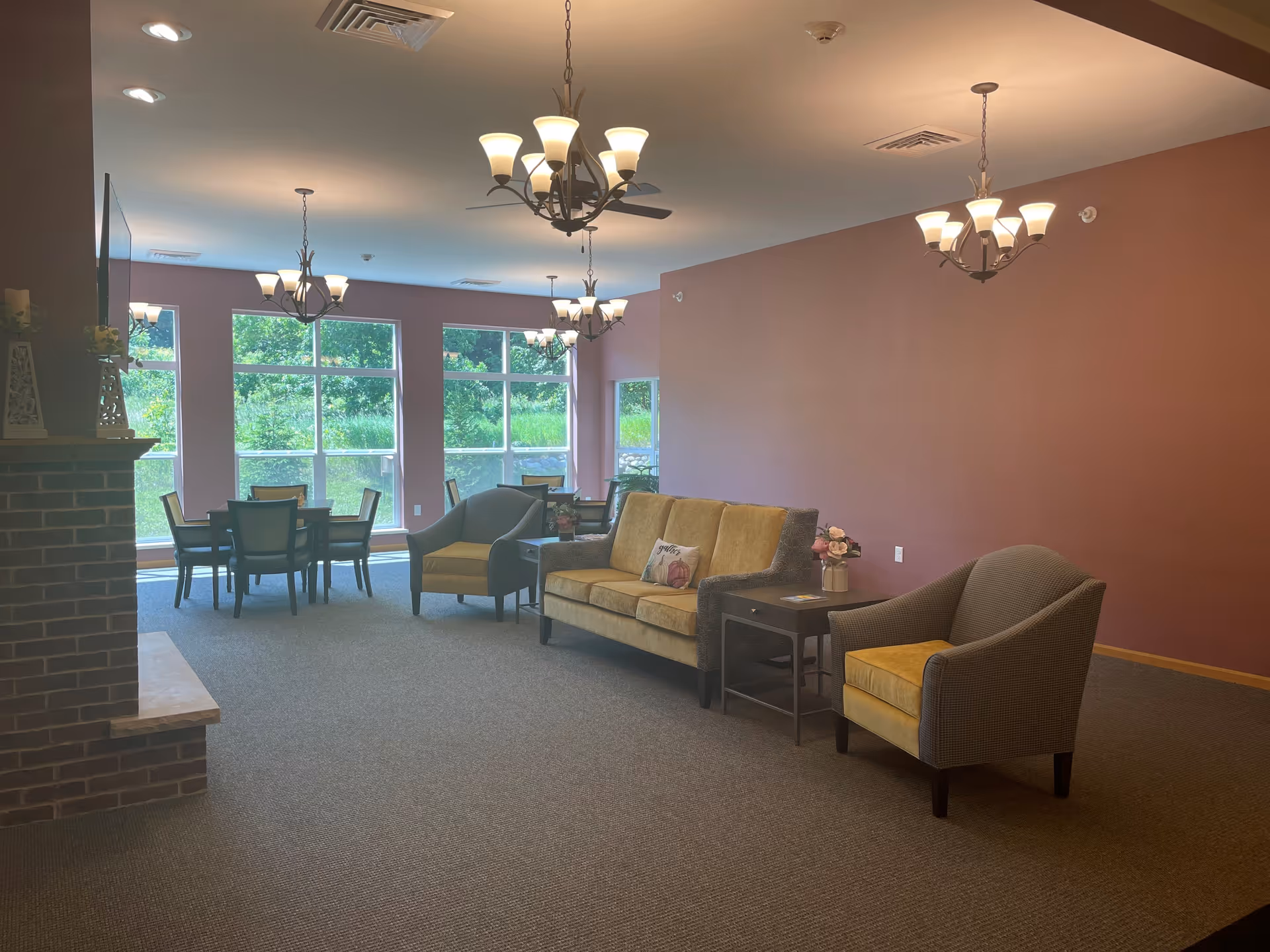 A senior living facility common area with a seating arrangement including a sofa and two armchairs with yellow cushions, side tables with flower vases, and several dining tables with chairs near large windows showing greenery outside. The room has multiple ceiling light fixtures and a brick fireplace on the left.