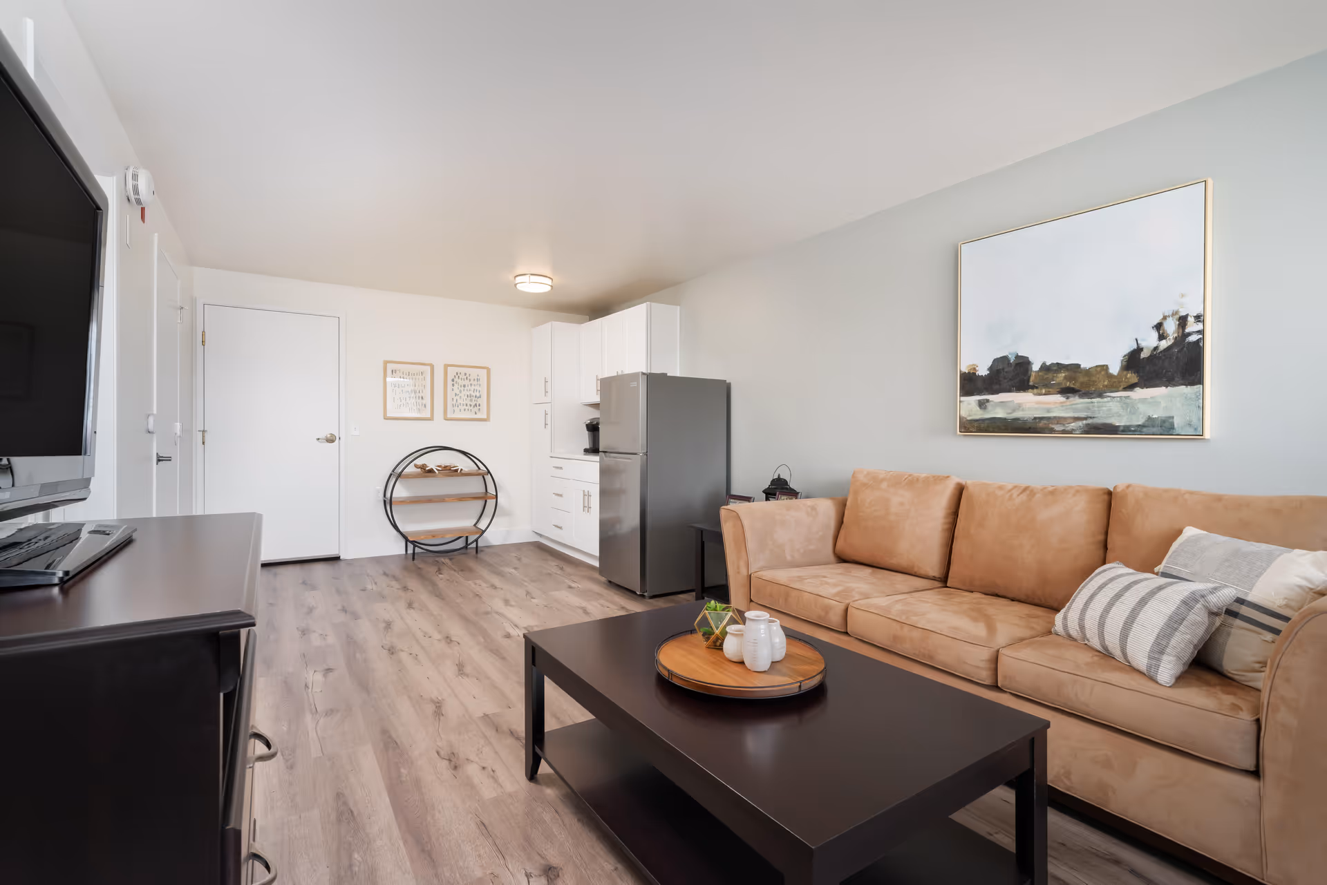 Modern living room with a tan sofa and striped pillows, dark coffee table, TV console, and a small kitchenette with a refrigerator.