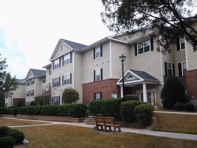 Exterior view of a three-story residential building with beige siding and red brick accents, surrounded by trimmed bushes and trees. A wooden bench is placed on a sidewalk in front of the building under a cloudy sky.