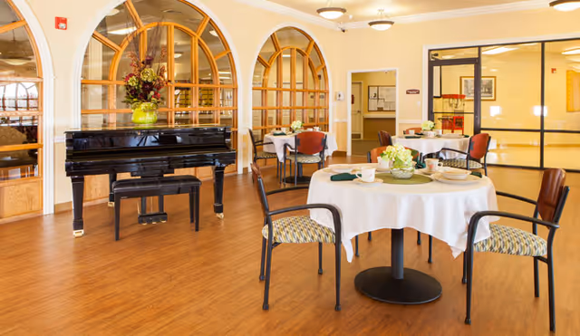 Dining room with round white‑clothed tables set for meals, chairs, and a black grand piano near arched interior windows.