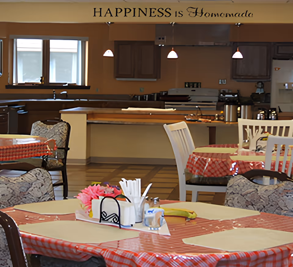 Interior view of a dining area with tables covered in red and white checkered tablecloths, set with placemats, napkins, and condiments. The background shows a kitchen area with cabinets, a stove, and hanging pendant lights. Above the kitchen area, a wall decal reads 'HAPPINESS is Homemade'.