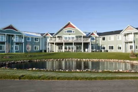 Front exterior view of a pastel-colored senior living building with a pond and lawn.