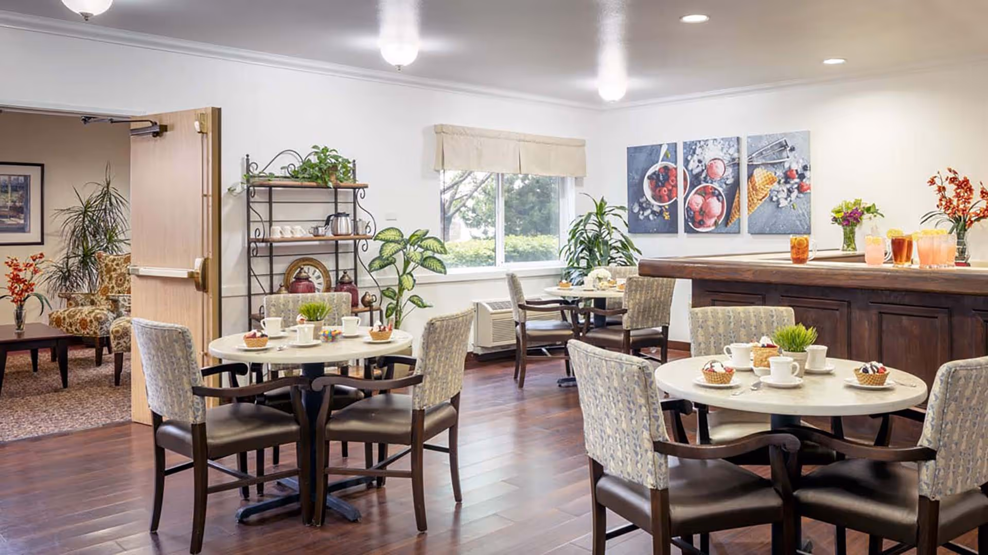 A bright dining area in a senior living facility with round tables set with cups, plates, and small desserts. The room features wooden flooring, upholstered chairs, a large window with a beige valance, and decorative plants. There is a wooden counter with glasses of beverages and colorful wall art depicting ice cream and berries. An open door leads to a cozy sitting area with patterned armchairs and a coffee table.
