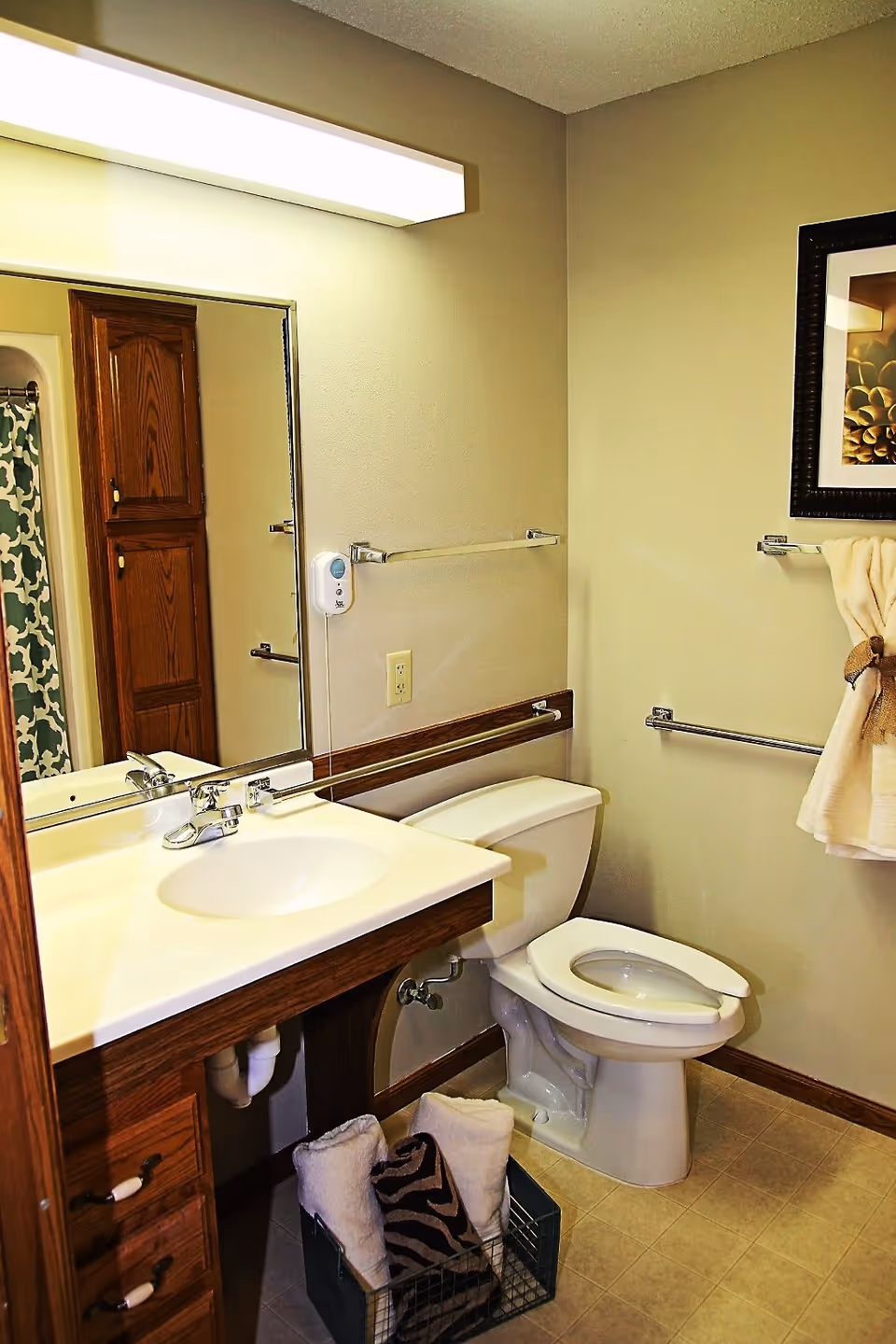 A bathroom with a white sink and countertop, a toilet with the lid up, and a large mirror above the sink. There is a wooden cabinet with drawers under the sink and a tall wooden cabinet reflected in the mirror. A towel rack with a beige towel tied with a brown ribbon is mounted on the wall next to the toilet. The floor is tiled, and a basket with rolled towels is placed under the sink. The walls are painted beige, and there is a framed picture hanging on the wall.