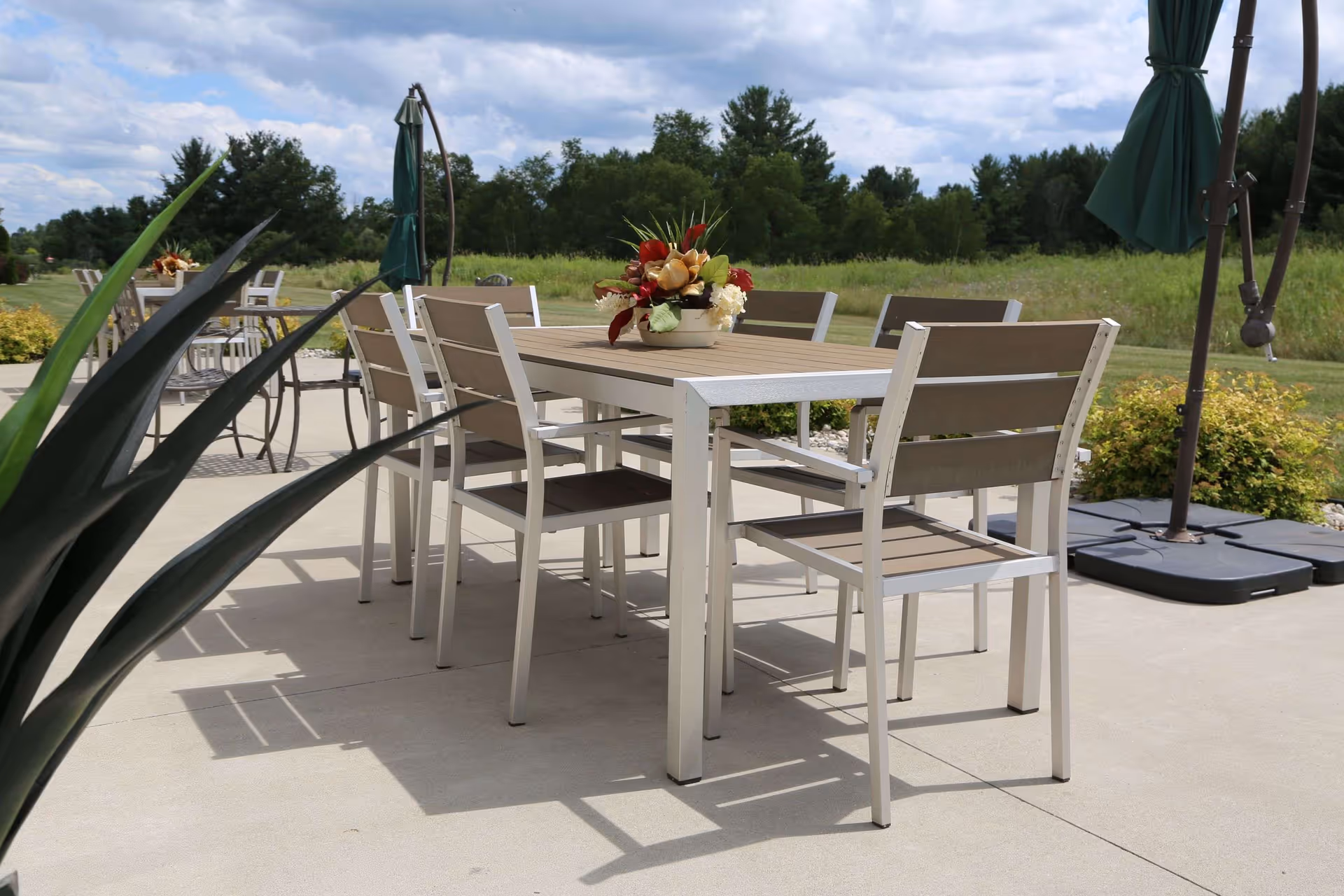 Outdoor patio area with a rectangular table and six chairs. The table has a floral centerpiece. There are green umbrellas and plants around the patio, with a grassy field and trees in the background under a partly cloudy sky.
