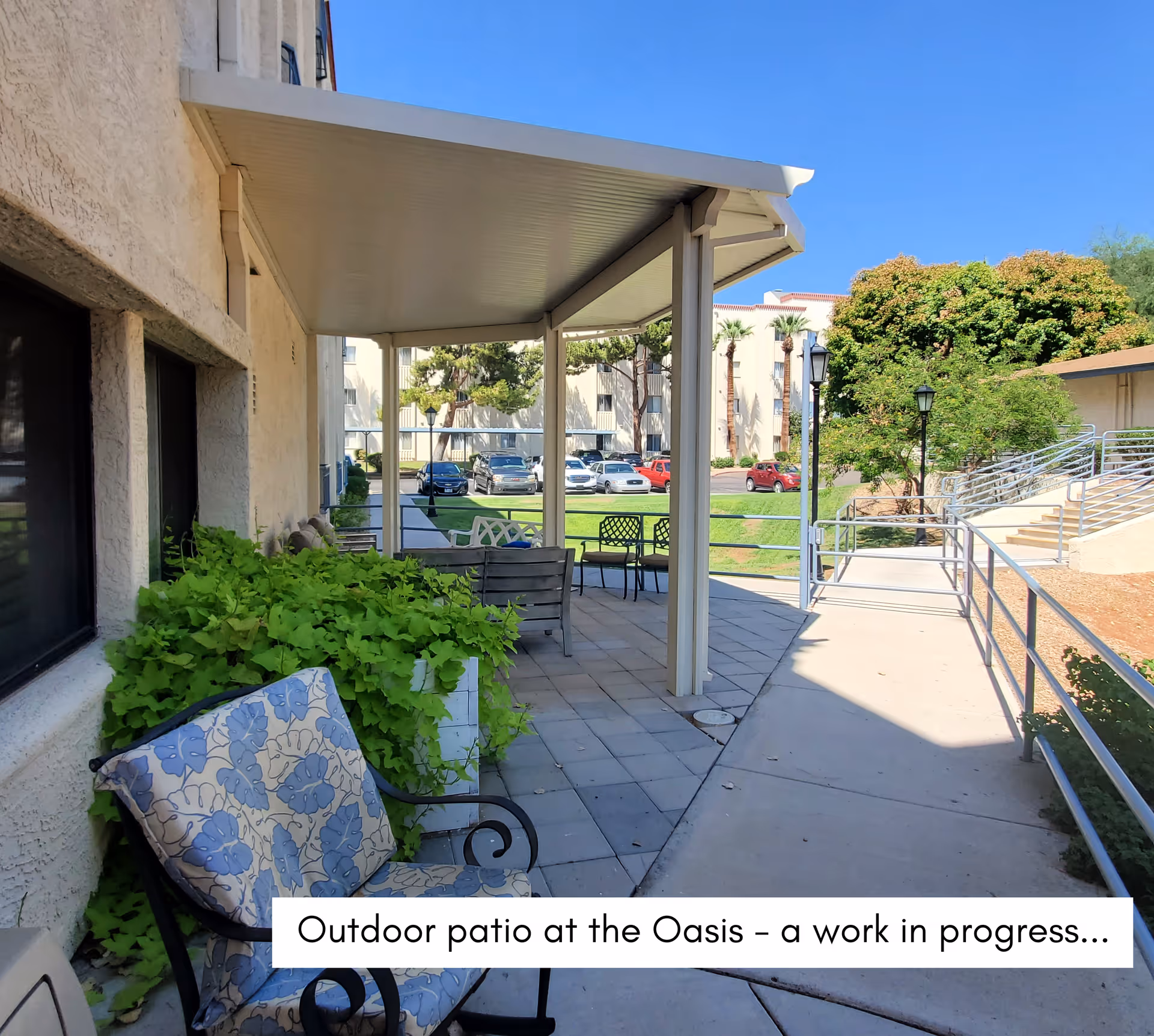 Covered outdoor patio with chairs and tables beside a walkway and landscaped courtyard at a senior living facility.
