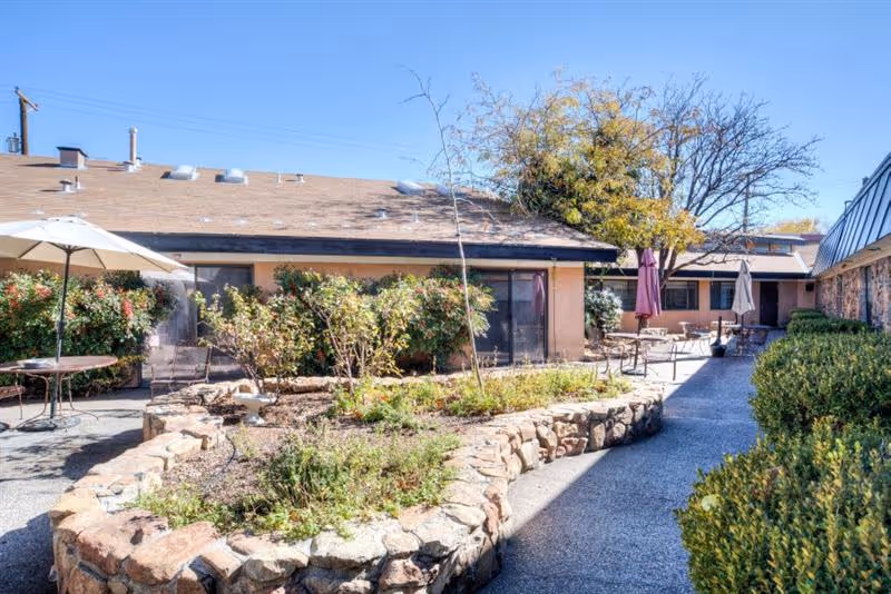 Outdoor courtyard area at Sandia Ridge Center with stone-bordered garden beds, patio tables with umbrellas, and surrounding single-story building under a clear blue sky.