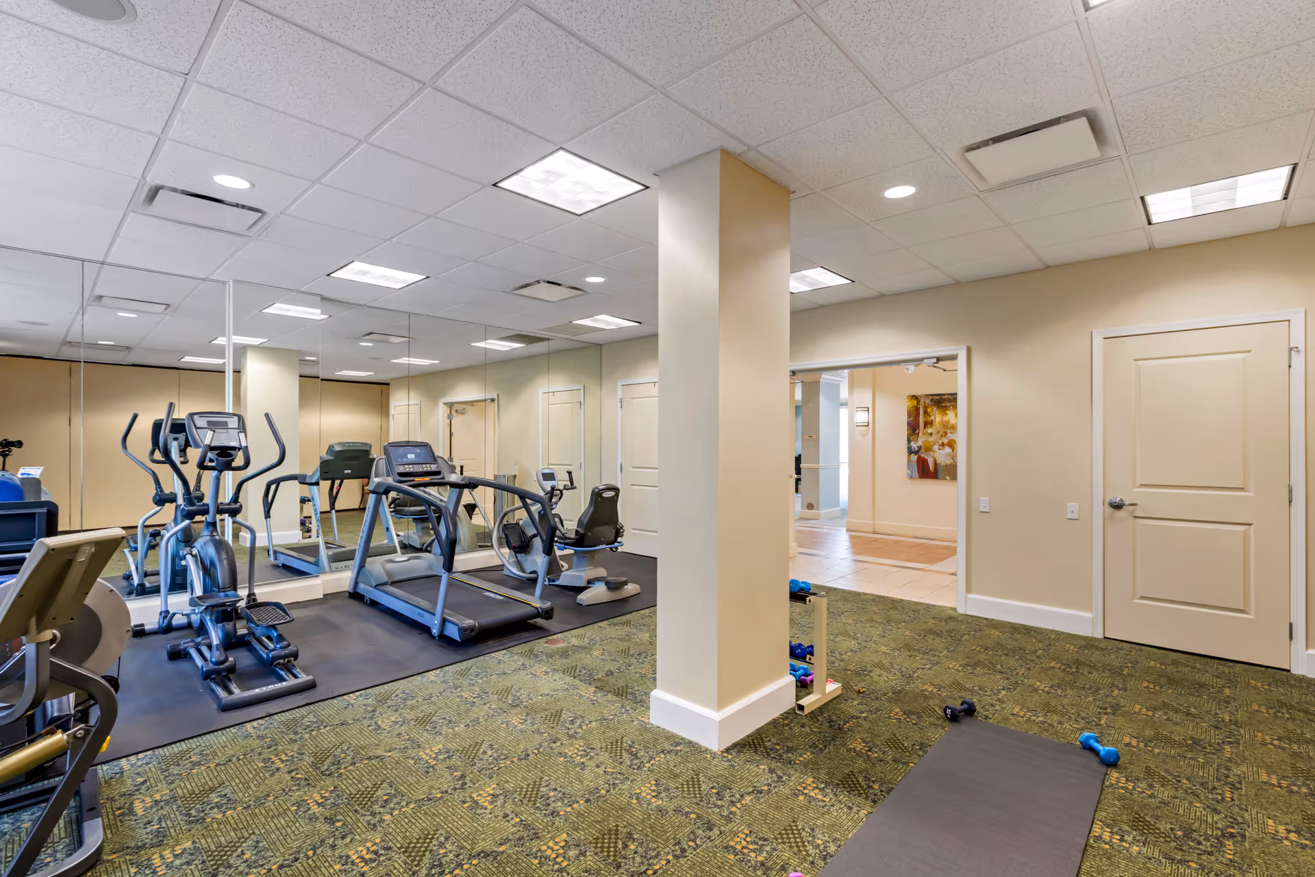 A fitness room with exercise equipment including a treadmill, elliptical machines, and a recumbent bike. The room has a green patterned carpet, beige walls, a mirrored wall, and a ceiling with recessed lighting. There are dumbbells and a yoga mat on the floor near a beige column. Two closed doors and an open doorway leading to a hallway are visible.