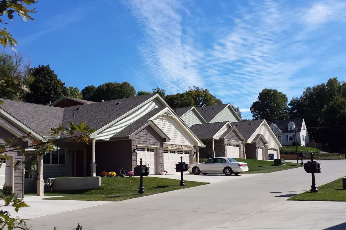 Row of single-story townhome-style units with attached garages, mailboxes, and a parked car along a paved driveway under a blue sky.