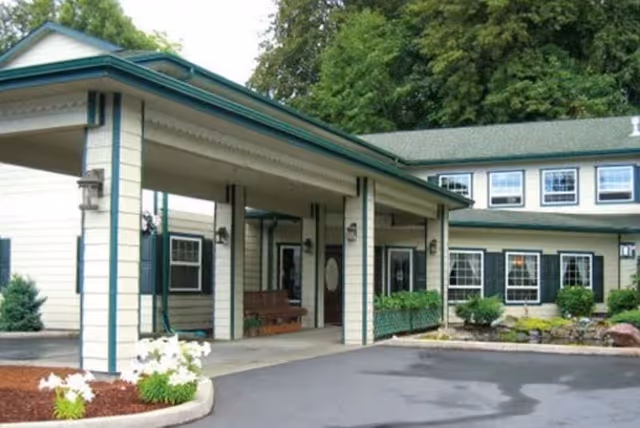 Exterior view of a senior living facility named The Amber, showing a covered entrance with columns, a bench, and landscaped greenery including bushes and flowers near the building.