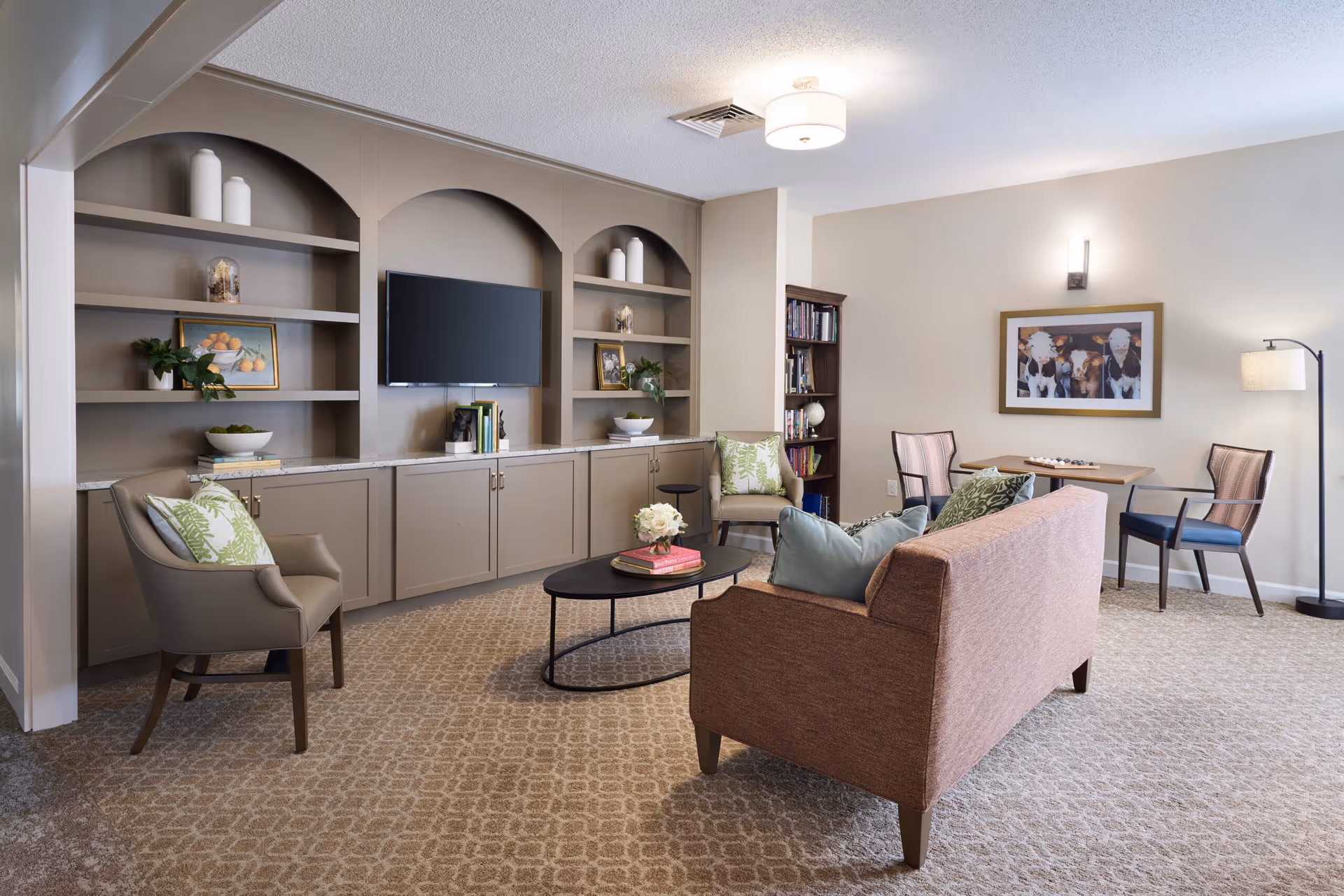 A cozy living room area in Addington Place of Lakeside Vista featuring a brown sofa with blue and green pillows, two armchairs with green patterned cushions, a black oval coffee table with books and flowers, built-in shelves with decorative items and a mounted flat-screen TV, a small table with two chairs, a floor lamp, and a framed picture on the wall.
