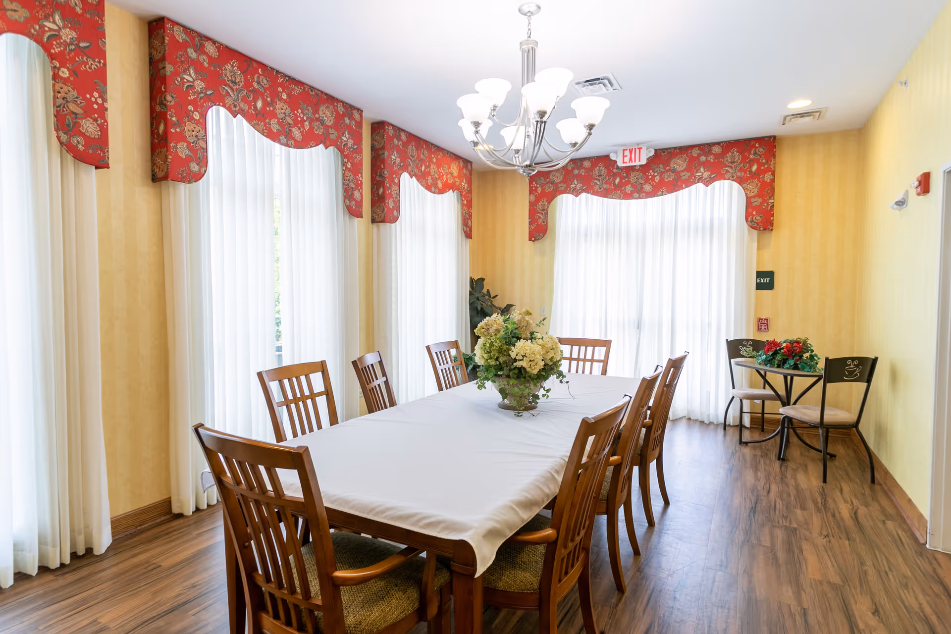 A dining room with a long rectangular table covered with a white tablecloth, surrounded by wooden chairs with cushioned seats. The room has large windows with sheer white curtains and red floral valances. A chandelier with multiple lights hangs above the table, and there is a floral centerpiece on the table. In the corner, there is a small round table with two chairs and a small flower arrangement. The floor is wood, and the walls are painted yellow with a subtle pattern.