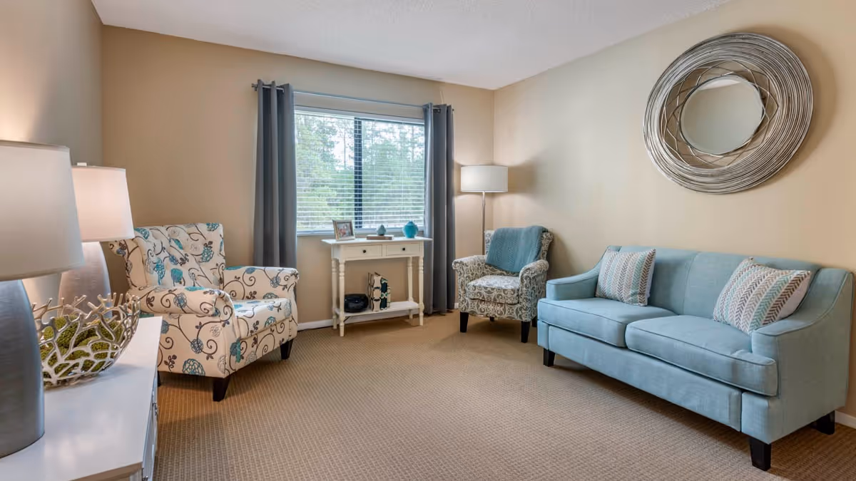 A cozy living room with a light blue sofa adorned with two patterned pillows, two armchairs with floral designs, a small white console table under a window with gray curtains, a floor lamp, and a decorative round mirror on the beige wall.