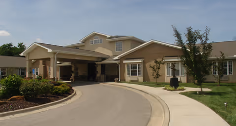 Front entrance of a beige senior living facility with a covered porte-cochère, curved driveway, sidewalk, and landscaped grounds under a blue sky.