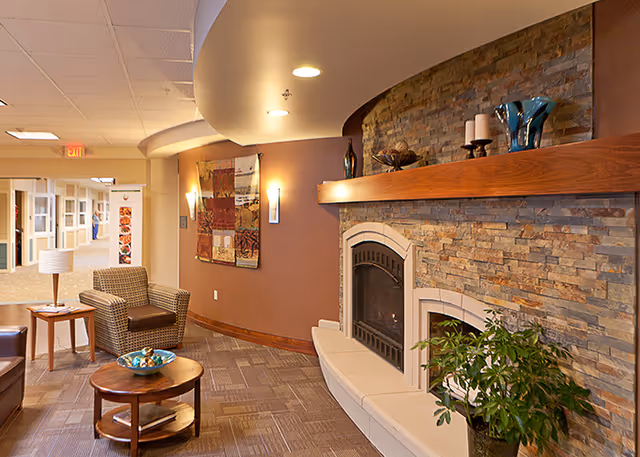 Interior view of a senior living facility lounge area featuring a stone fireplace with a wooden mantel decorated with candles and vases. There is a comfortable armchair and a small round wooden table with decorative items. The hallway extends into the background with additional seating and wall decorations.