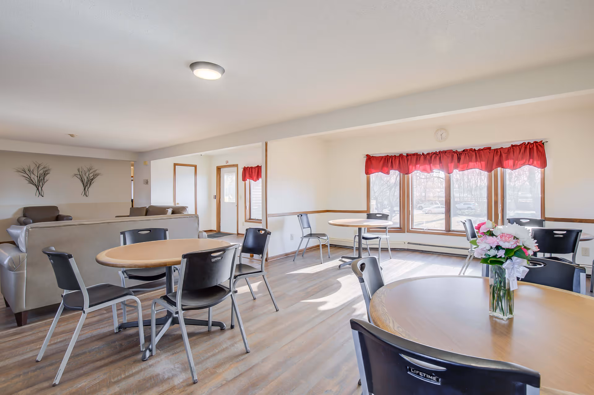A bright and spacious common area in a senior living facility with multiple round tables and black chairs arranged around them. There are beige sofas in the background and large windows with red valance curtains letting in natural light. A vase with pink and white flowers is placed on the nearest table.
