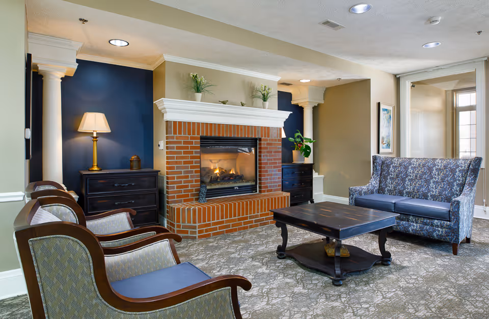 A cozy senior living facility common area with a brick fireplace in the center, flanked by two white columns and dark blue walls. There are two wooden armchairs with patterned cushions on the left, a dark wooden coffee table in the middle, and a blue patterned loveseat on the right. The room is softly lit with a table lamp on a black chest of drawers and natural light coming through a window on the far right.