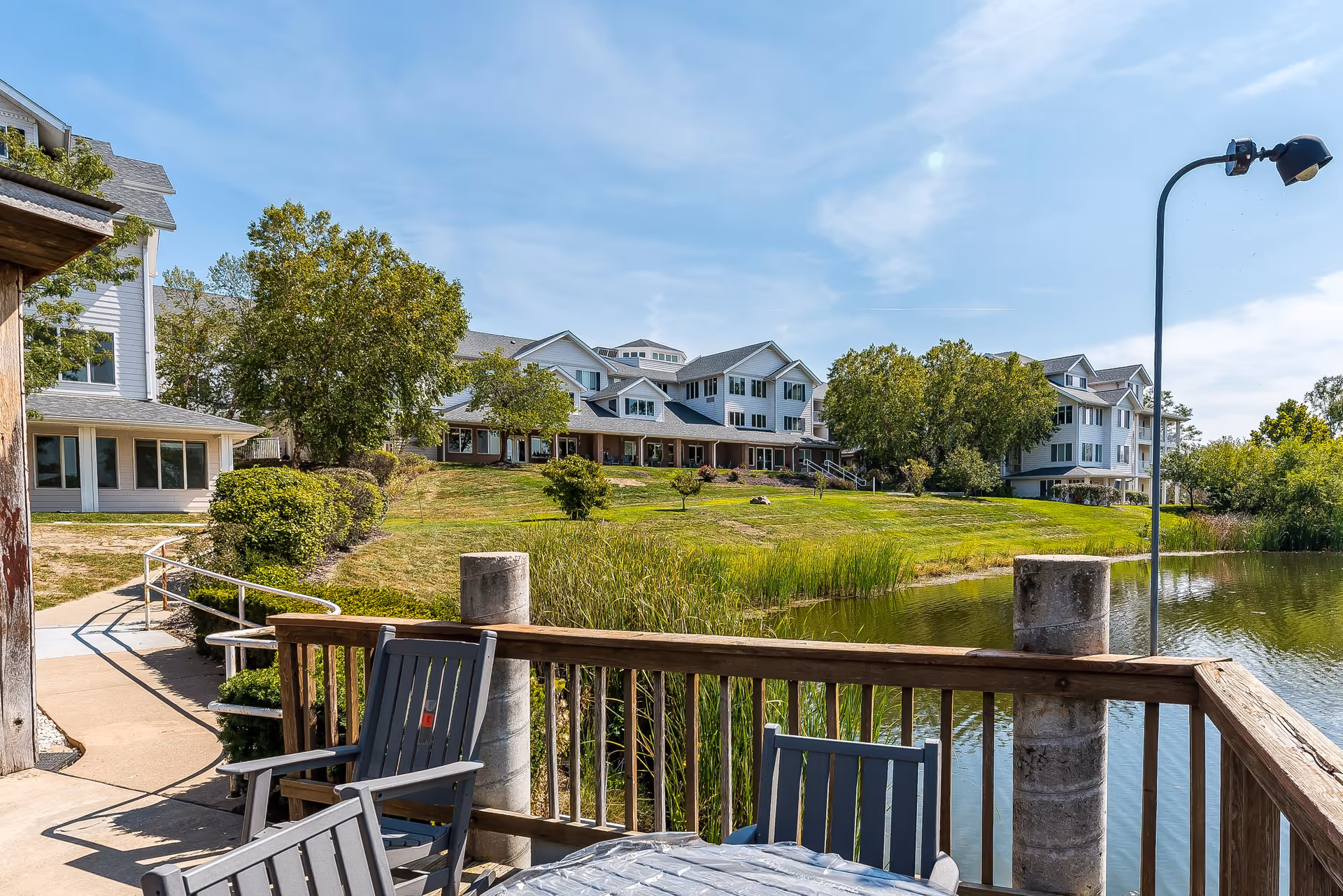 View of a senior living facility building named Solstice Senior Living at Columbia from a wooden deck overlooking a pond. The deck has several gray chairs and a table. The building is surrounded by green grass, trees, and a clear blue sky.